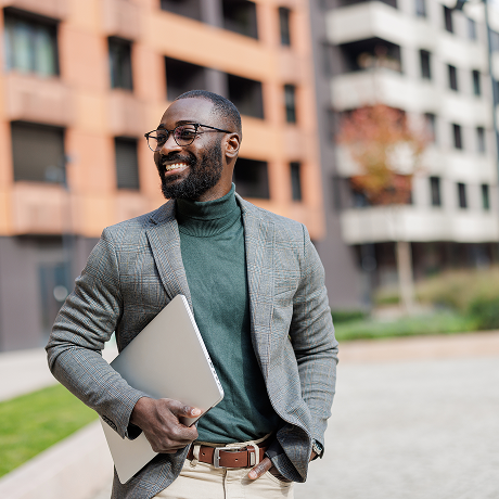 Man in suit stands before a number of buildings with a laptop in hand